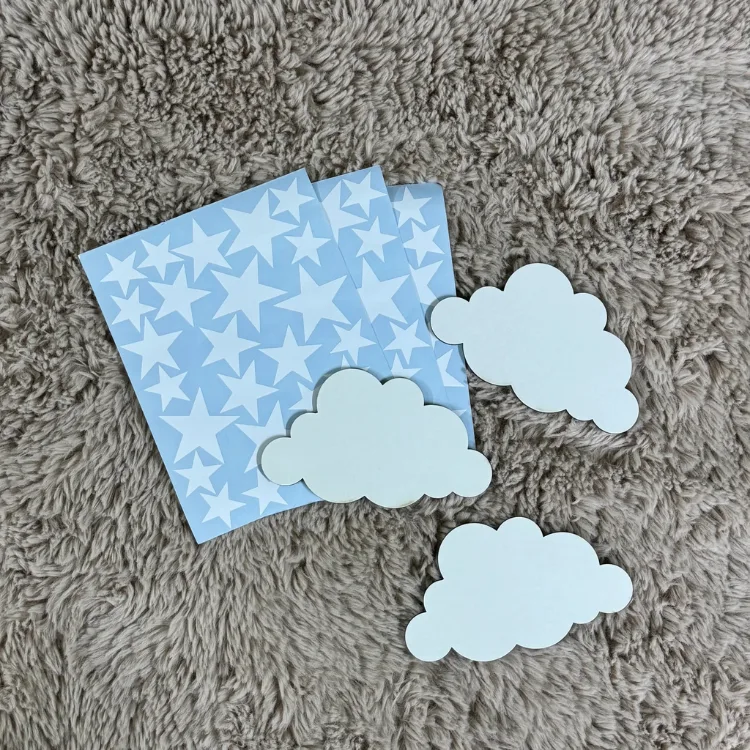 Bulut ve yıldız duvar süsü setinde beyaz yıldız sticker sayfaları ve krem rengi üç bulut, tüylü halı üzerinde