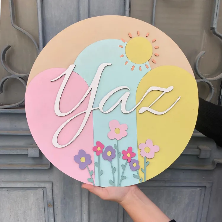 Bahar temalı kapı süsünün yakın planı; pastel renkli yuvarlak zeminde güneş, mor ve pembe çiçekler, beyaz örnek isim