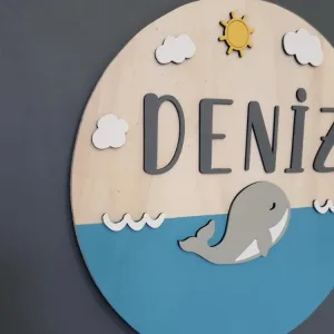 Deniz kapı süsü yandan; kabartma gri balina, sarı güneş, beyaz bulutlar ve antrasit harflerin katmanlı görünümü