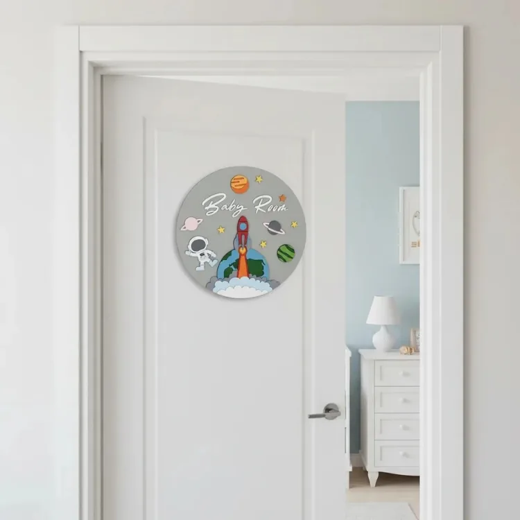 İsimli uzay temalı kapı süsü beyaz kapıda; gri zeminde Baby Room yazısı, roket, astronot, gezegen ve yıldız figürleri