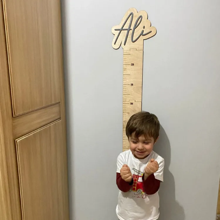 İsme özel ahşap boy cetveli duvarda, önünde gülümseyen bir çocukla; üstte örnek isim ve santimetre çizgileri