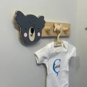 Koala askısı yandan; katmanlı antrasit koala yüzü, pembe yanaklar ve askı çubuğunda asılı bebek kıyafeti
